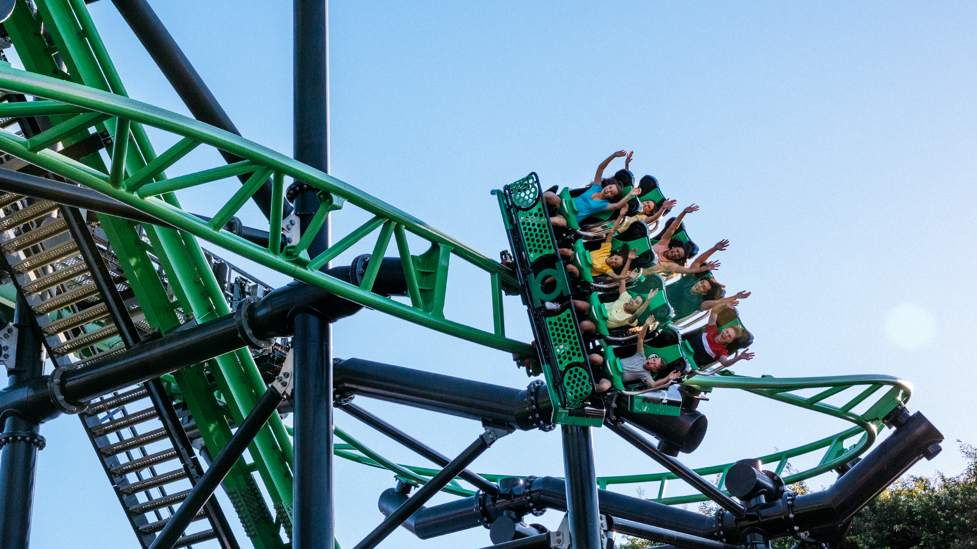 A group of people ride a green and black roller coaster with their arms raised, against a clear blue sky background.