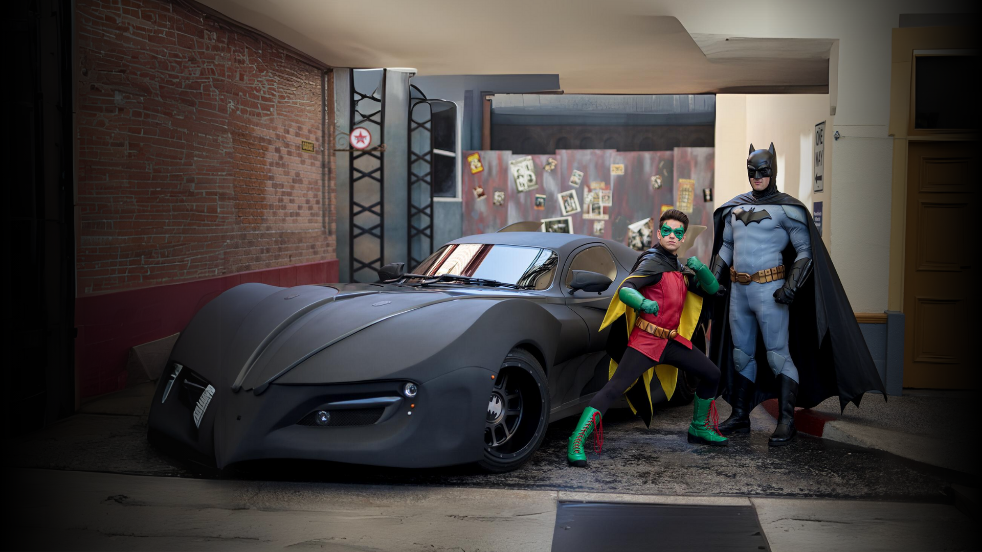 Two people dressed as Batman and Robin pose next to a black, custom-designed Batmobile in an indoor setting with posters and a brick wall in the background.